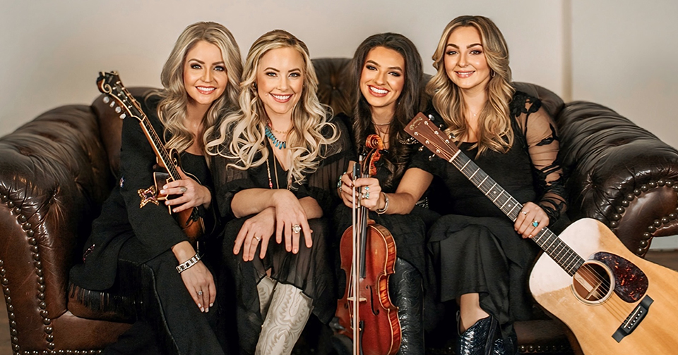 Photo of four women holding instruments and sitting on a brown leather sofa