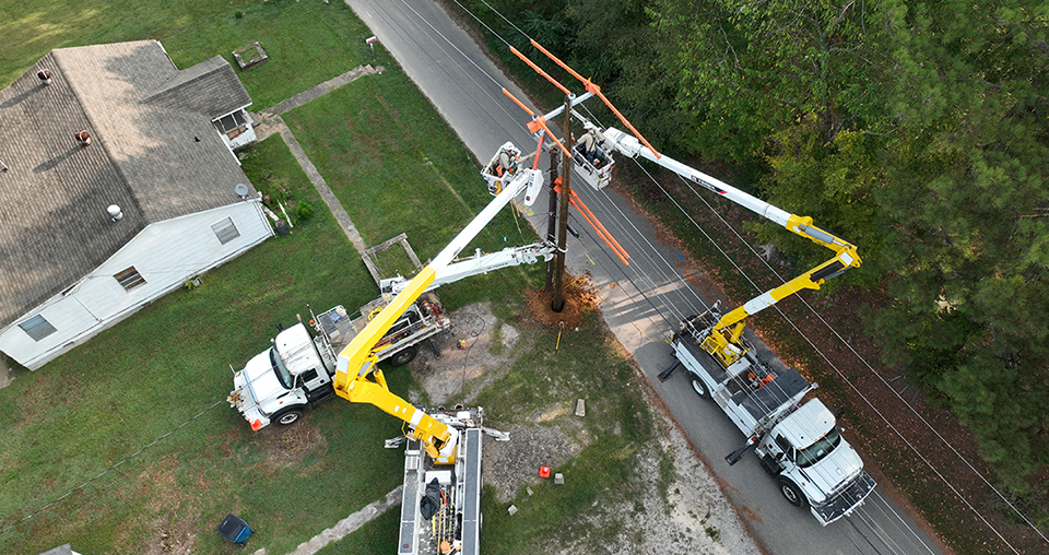 Photo showing three bucket trucks. Two bucket trucks have the buckets extended, with one lineworker in each bucket, working to change the pole on a main distribution line.