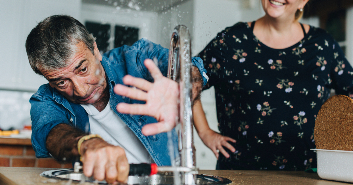 Photograph of a man in his mid-50s failing to fix a faucet in the kitchen sink and trying to prevent water from splashing on him