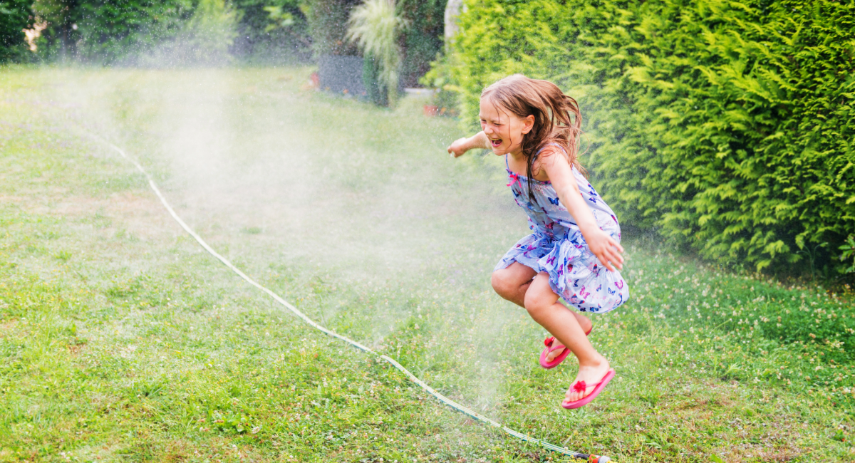 Photo of a young girl, probably around 6 or 7 years old, jumping through a sprinkler