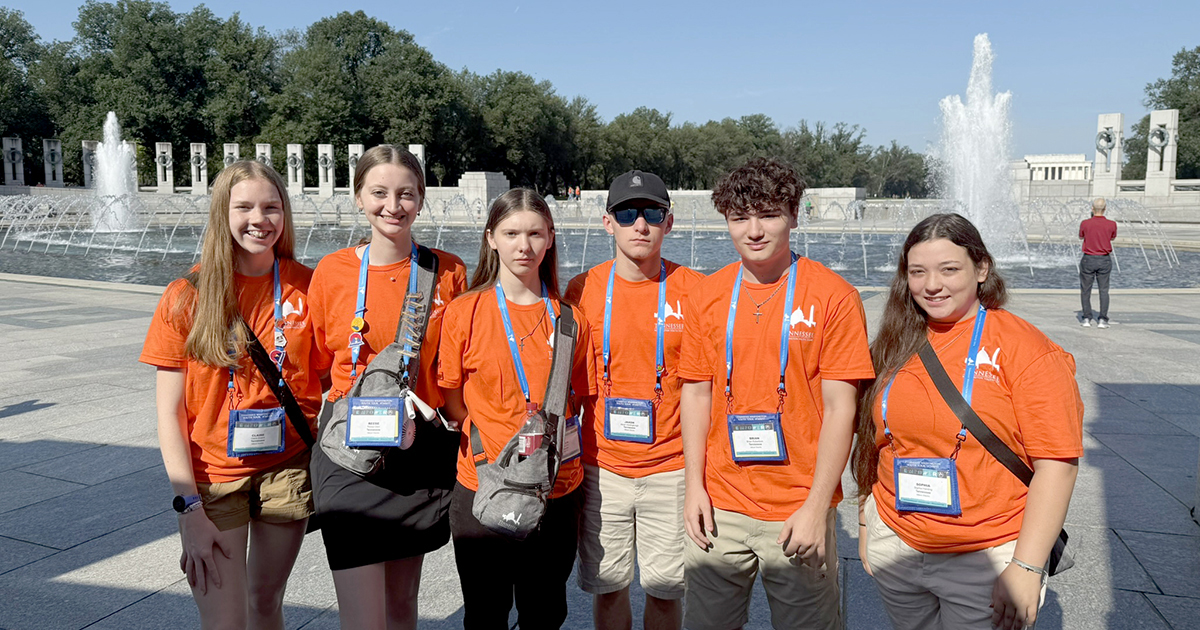 A group of students wearing matching orange T-shirts and blue lanyards with name tags stand in front of the World War II Memorial on the National Mall in Washington, D.C., with a large fountain, trees, and columns in the background