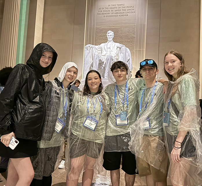 A group of students wearing raincoats and ponchos stand in the Lincoln Memorial with the statue of President Lincoln sitting on a chair in the background