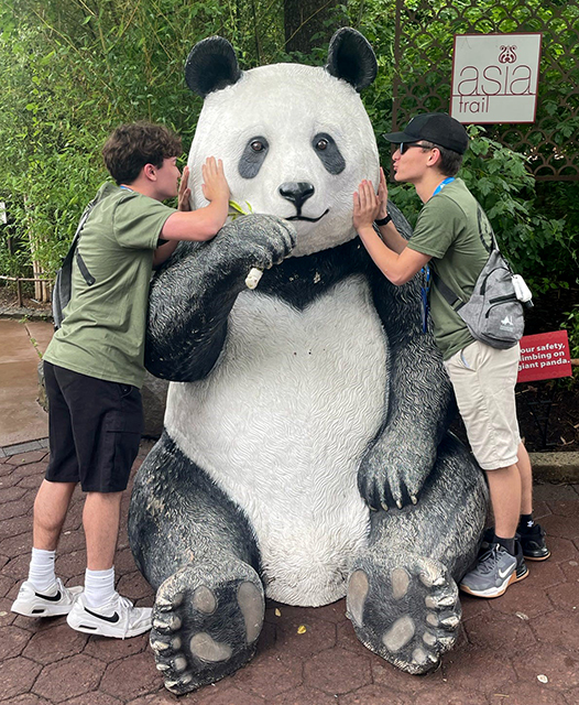 Two students wearing matching sage T-shirts flank a large panda statue, pretending to kiss the panda on the cheek, at the National Zoo in Washington, D.C.
