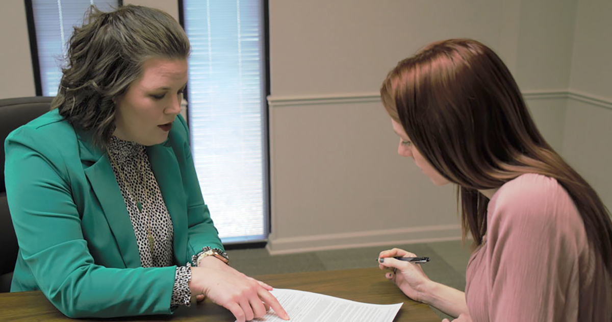 Photo showing two women sitting across from one another at a desk. The woman on the left is pointing to place on the paper on the desk between them where the woman on the right needs to sign a contract.