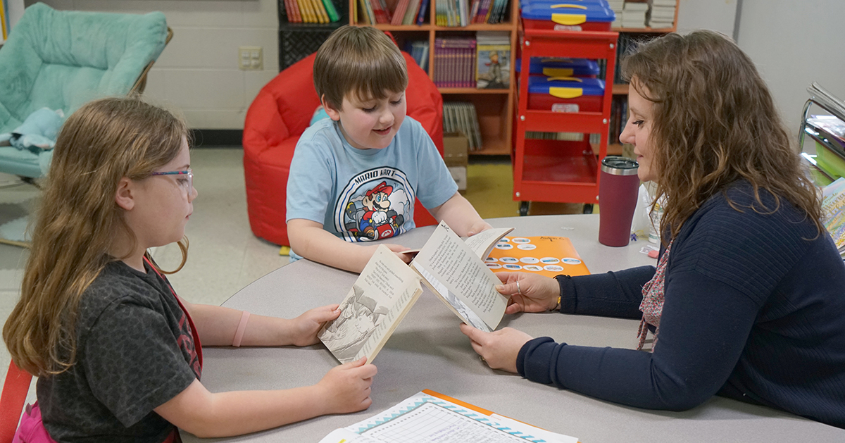 Photo of two young students reading with their teacher. The students and teacher are each holding a book.