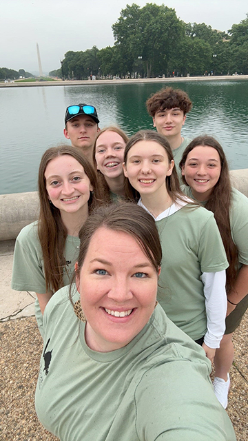 Chaperone and students wearing matching sage T-shirts take a selfie photo with the reflecting pool and Washington Monument behind them