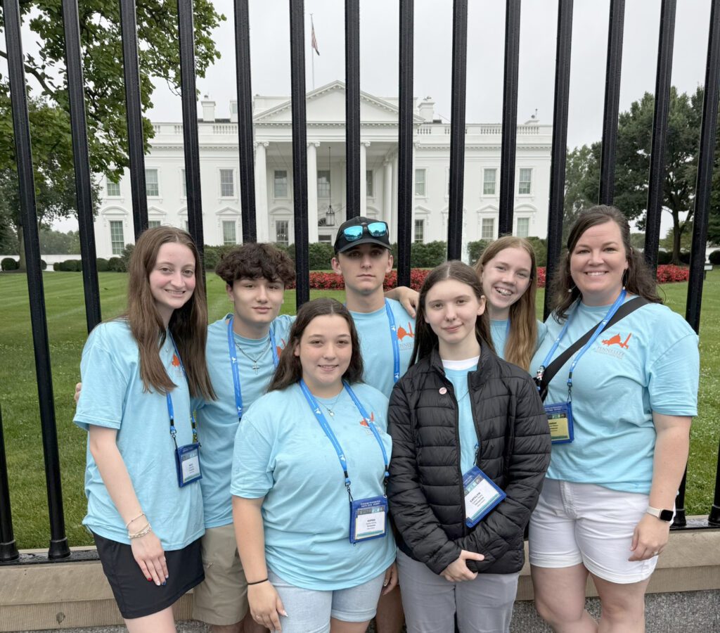 A group of students wearing matching blue T-shirts and blue lanyards with name tags stand in front of the White  House in Washington, D.C.