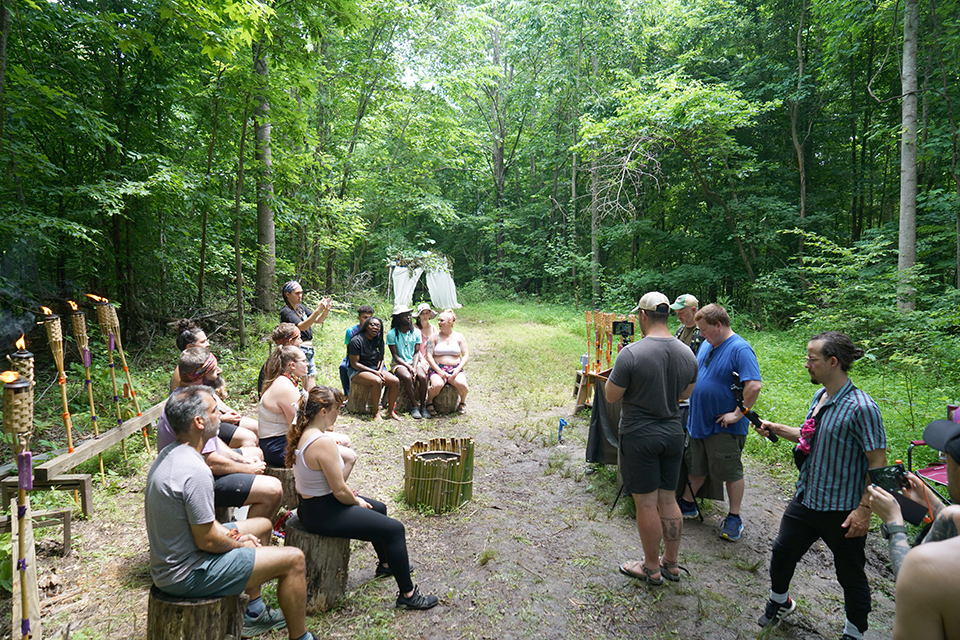 A group of people gathered in a clearing in the woods, with some seated on wooden logs arranged in a semi-circle and others standing. Tiki torches line the left side of the clearing, and a small bamboo structure stands in the center. The background is filled with dense green foliage and trees.