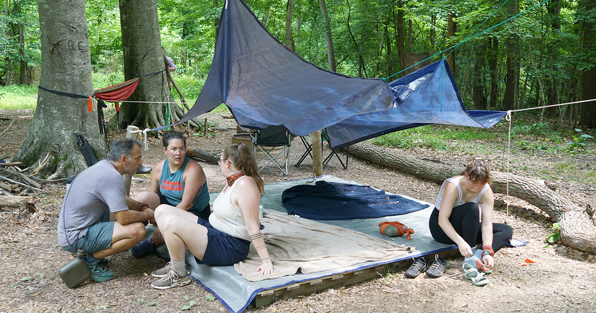 A group of four people are camping in the woods. Three are seated on a tarp-covered platform under a blue tarp canopy, while one person sits on the ground adjusting their shoes. The campsite is surrounded by trees, with a hammock tied between two trees in the background. Camping gear and supplies are scattered around the area.