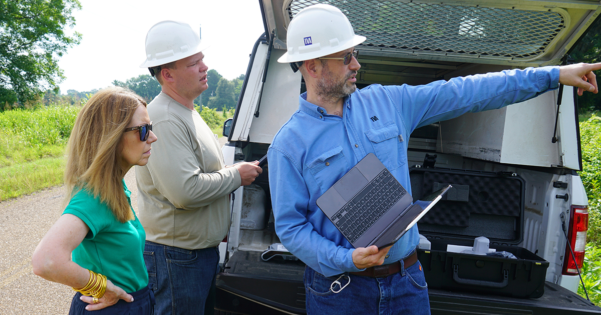 Two men in hard hats and a woman stand near a utility vehicle as one man points into the distance during a drone inspection.