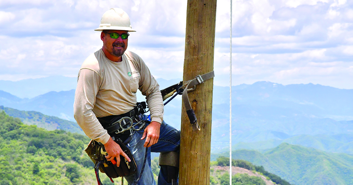 Donny Thomas, a lineworker, wearing a safety harness and helmet is secured to a wooden utility pole. The background shows a scenic view of mountains and cloudy skies in Guatemala.