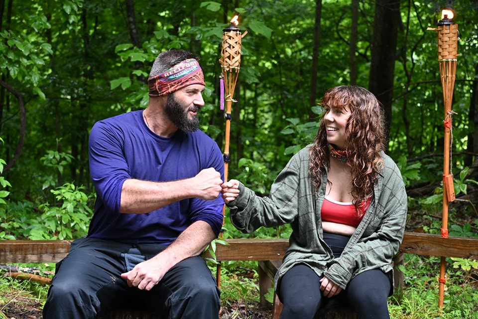 Two people sit on a wooden bench in the woods, fist-bumping each other while holding torches. The background is filled with green foliage and trees.