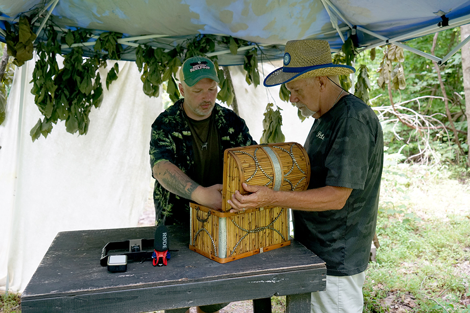 Two individuals stand under a canopy with hanging leaves, focused on a wooden chest placed on a table in front of them. One wears a green cap and has tattoos on their arm, while the other wears a straw hat and a dark shirt. 