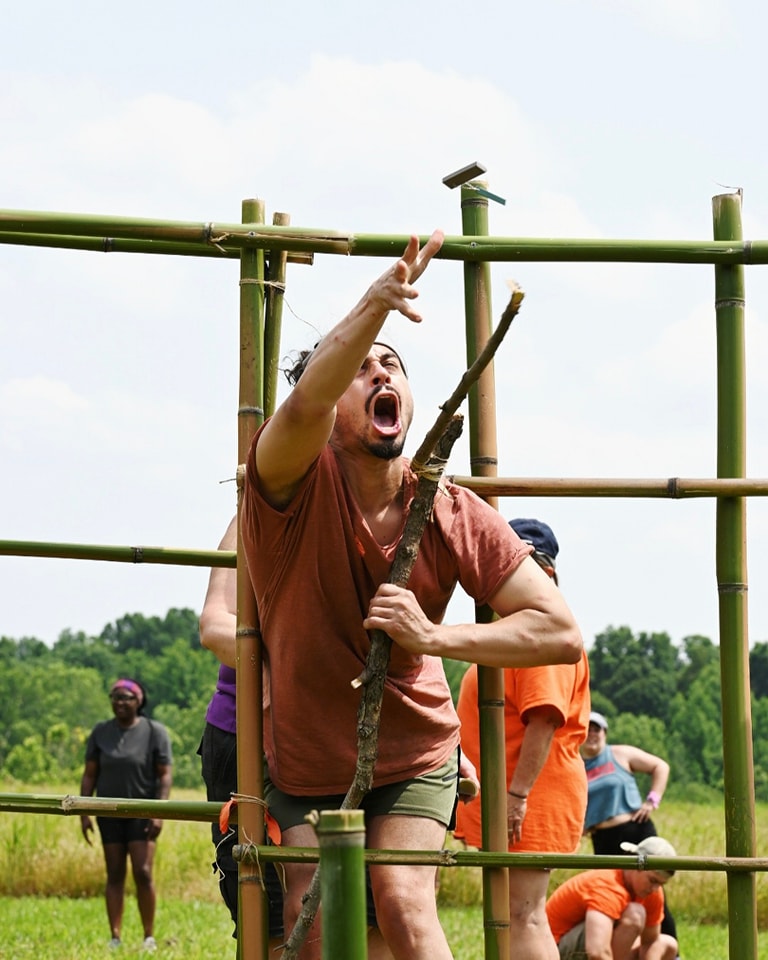 A participant in the game 'Surviving Reelfoot' reaches upward with a stick toward a piece of flint, the reward for completing a challenge involving bamboo structures. Other participants in orange shirts are visible in the background, engaged in similar activities. The scene takes place on a grassy field with trees in the distance under a partly cloudy sky.