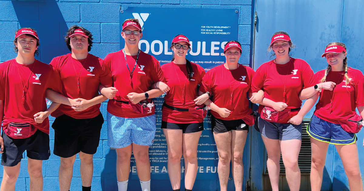 Seven teenage lifeguards from the Northwest Gibson County YMCA stand in a line with arms linked, wearing red shirts and red visors labeled "GUARD." They smile at the camera in front of a blue wall displaying a "POOL RULES" sign.
