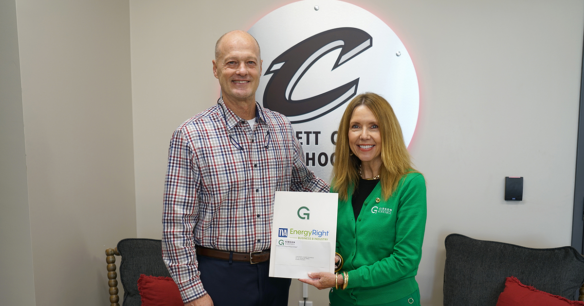 Two people stand in front of a wall with a large "C" logo. The man wears a checkered shirt and dark pants; the woman wears a green sweater and holds a document labeled "TVA EnergyRight." Both are smiling at the camera.