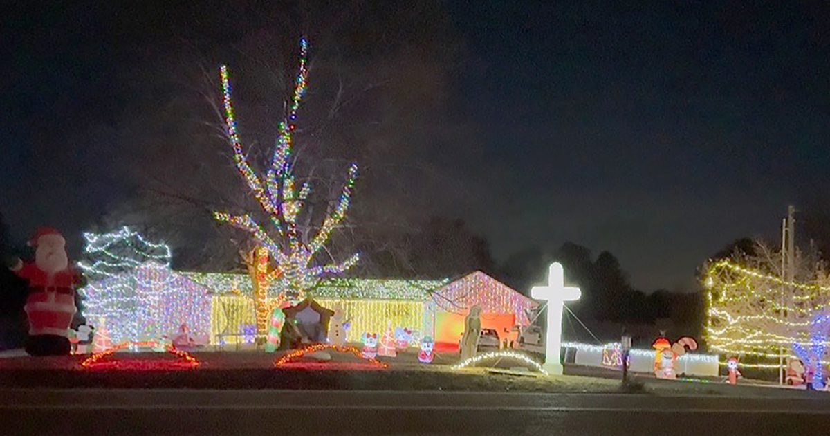 A single-story house and yard decorated with elaborate Christmas lights and inflatables at night. The house is outlined in white lights, with the roof covered in dense strands of multicolored lights. A large leafless tree in the front yard is wrapped in multicolored lights, and another tree on the right is wrapped in yellow lights. Inflatables include a large Santa Claus on the left, snowmen, and other holiday figures scattered across the yard. A bright white illuminated cross stands prominently near the center-right of the display. The entire scene glows against a dark night sky.