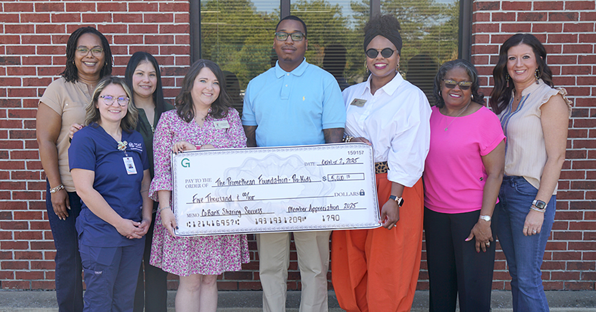 A group of eight people standing in front of a red brick wall holding an oversized check. The check is made out to The Promethean Foundation, Inc.” for $5,000, with the memo “Member Appreciation 2025.