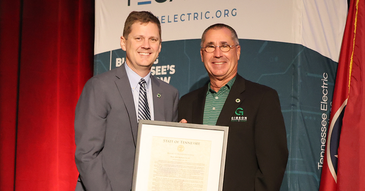 Two individuals stand side by side on a stage holding a framed Joint House Resolution. One person is wearing a gray suit with a striped tie, and the other is wearing a dark blazer with a green collared shirt featuring the Gibson Electric logo. Behind them is a banner with text that includes “Tennessee Electric Cooperative Association," along with a red curtain and part of a Tennessee state flag.