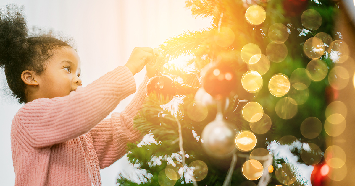 Child wearing a pink sweater decorates a Christmas tree with ornaments, surrounded by warm glowing lights and festive greenery.