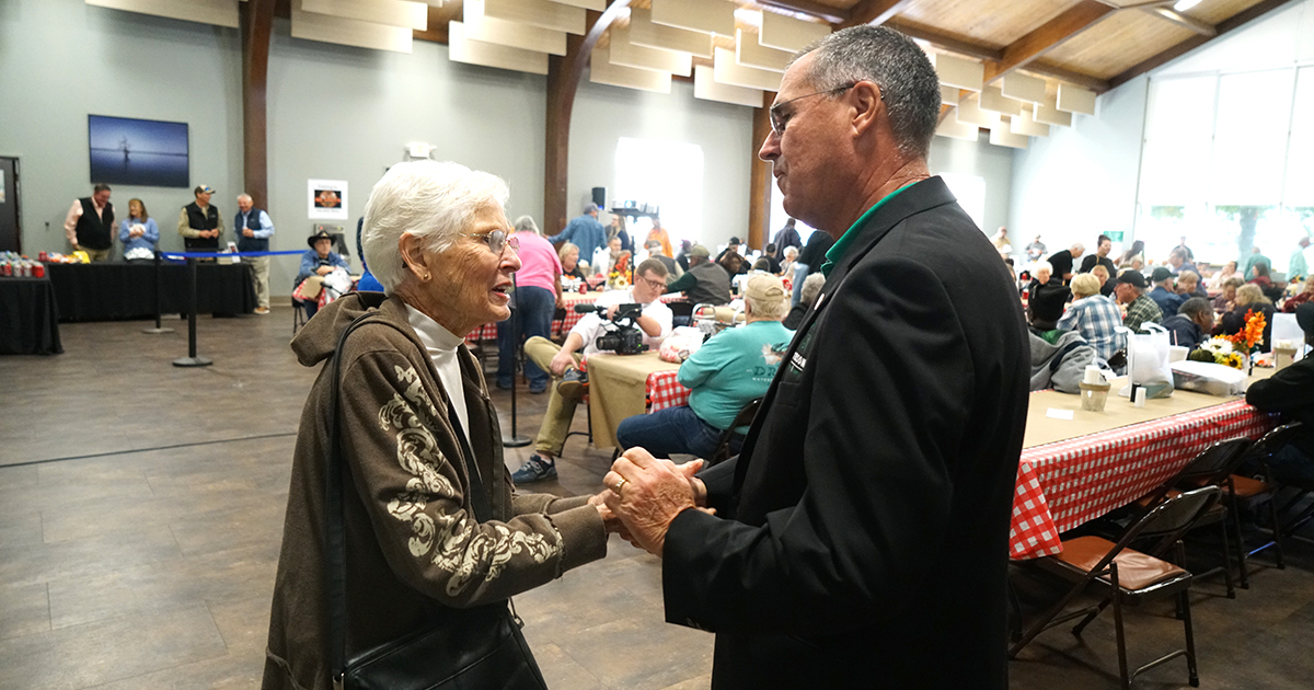 An older woman and a man shake hands while speaking inside a large community hall filled with people seated at long tables. Attendees visit, eat, and mingle in the background beneath high wooden ceilings, with a casual, welcoming event atmosphere.