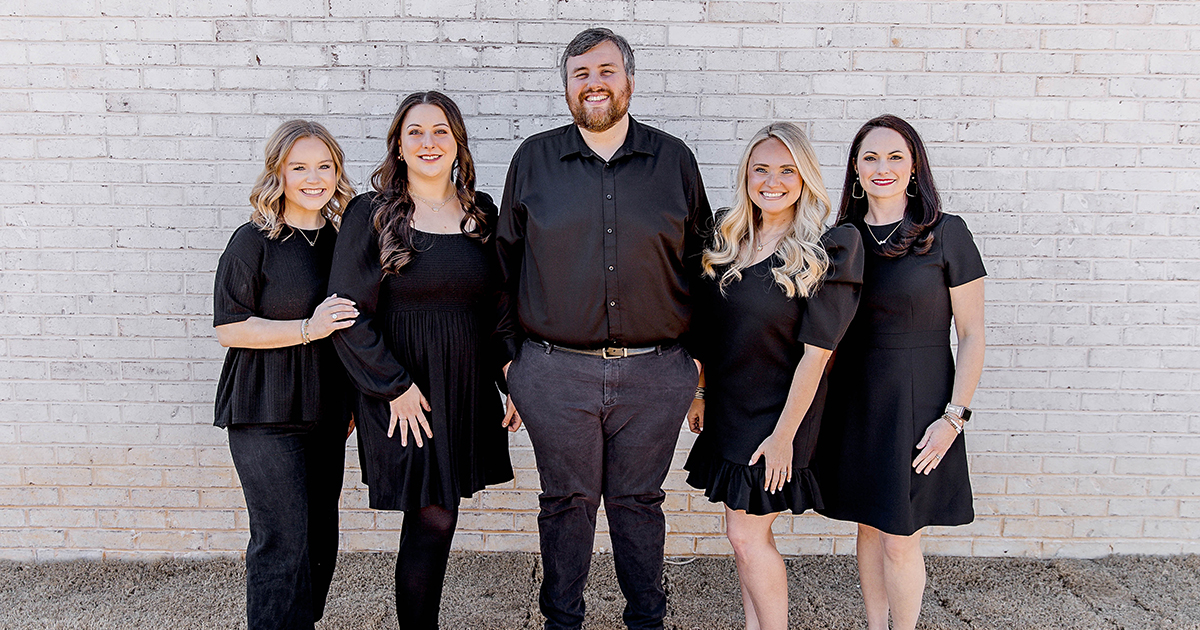 Five adults stand side by side against a light brick wall, dressed in coordinated black outfits. One person stands in the center wearing a black button-down shirt and pants, with two people on each side wearing black dresses or tops, posed closely together on a concrete surface.