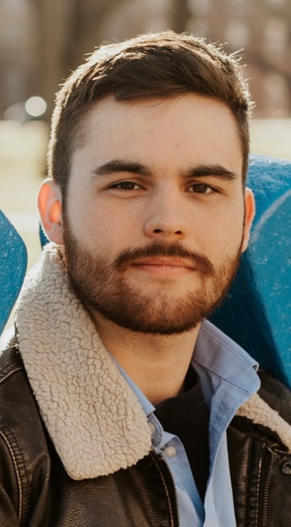 Close-up portrait of a young man outdoors, wearing a brown jacket with a shearling collar over a light blue button-down shirt, with trees and soft sunlight in the background.