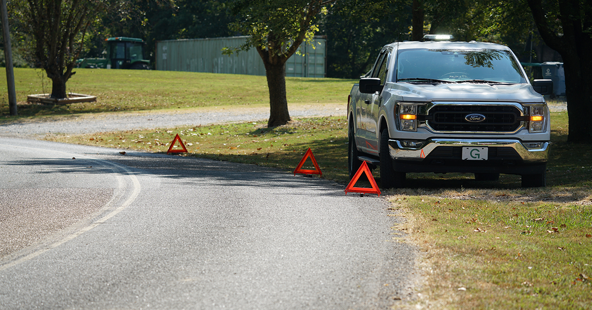 A white pickup truck is parked along a rural roadside, with three triangular emergency warning reflectors placed on the pavement behind it. Trees, grass, and a lightly traveled road are visible in the background on a clear day.