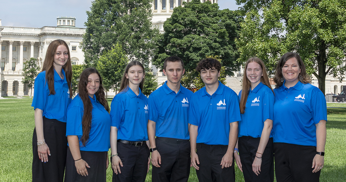 Seven people wearing matching blue polo shirts stand side by side on a grassy lawn, with the U.S. Capitol and leafy trees visible in the background on a sunny day.