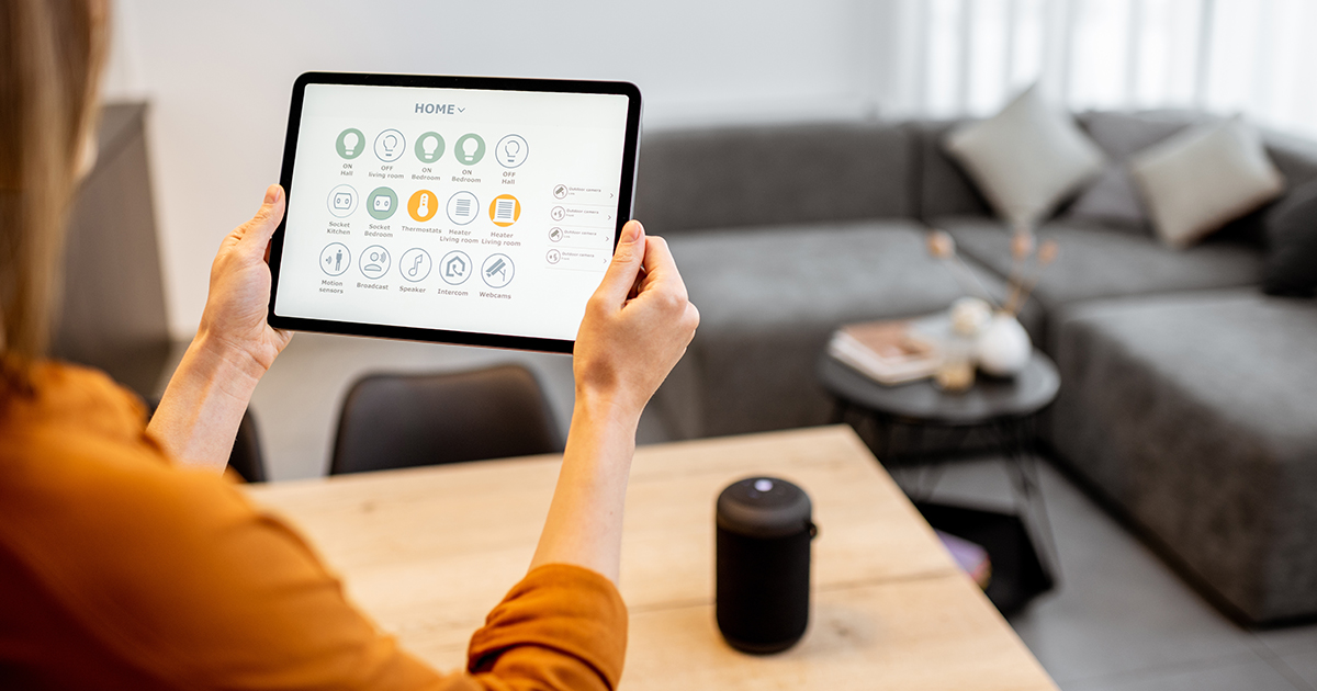 A person seated at a table holds a tablet displaying a smart‑home control interface. A small cylindrical smart speaker sits on the table in front of the person. In the background is a modern living room with a large gray sectional sofa, a round side table, and soft lighting.