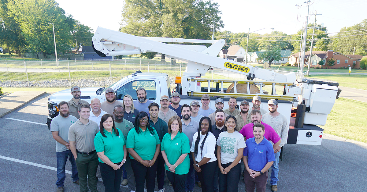 A large group of fiber services employees stands in front of a Gibson Connect bucket truck in a parking lot, with trees and houses visible in the background.