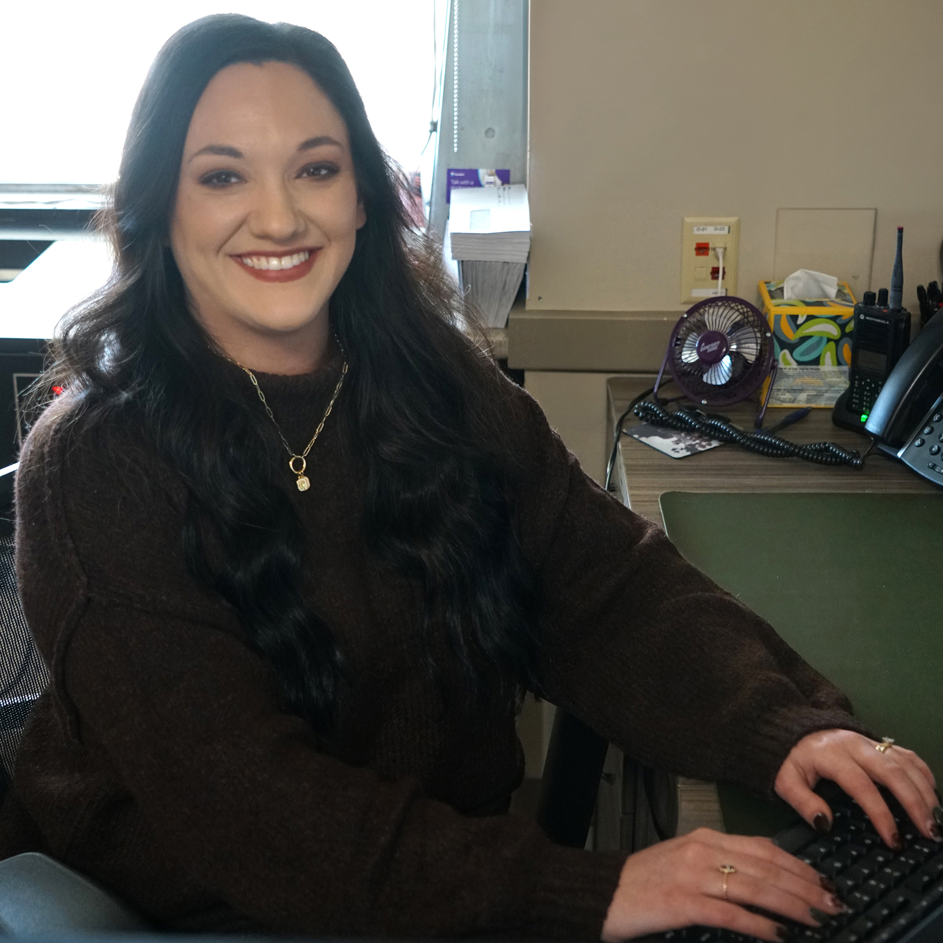 Smiling woman seated at an office desk, typing on a keyboard, with a telephone, small desk fan, and office supplies nearby.