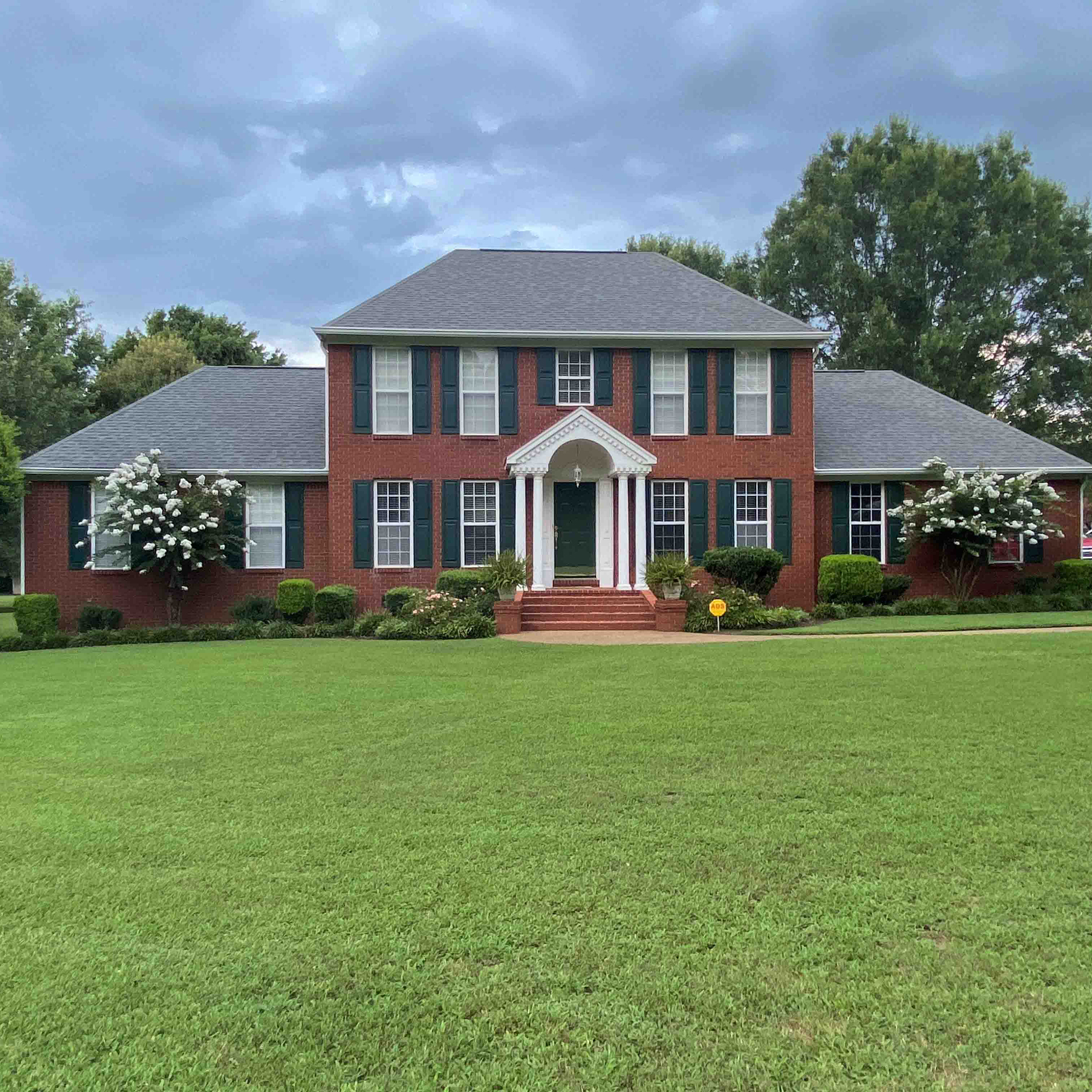 Large two-story red brick building with a centered front entrance, white columns, black shutters, and a manicured lawn in the foreground.