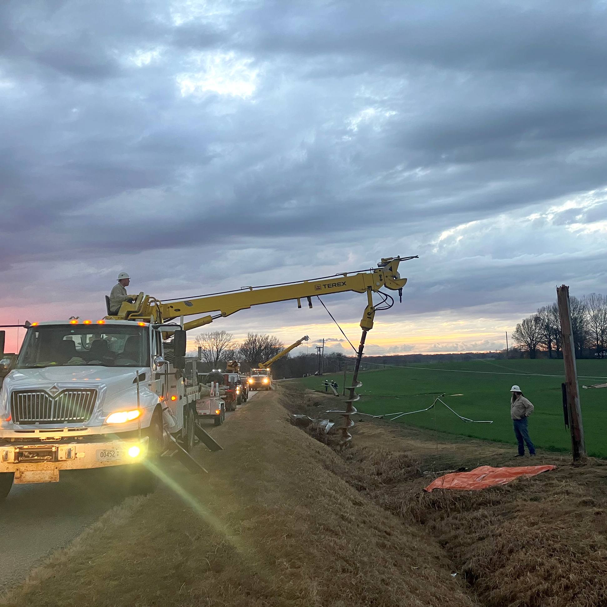 Utility crew using a bucket truck and equipment to install power poles along a rural roadside at dusk, with trucks lined up in the background.