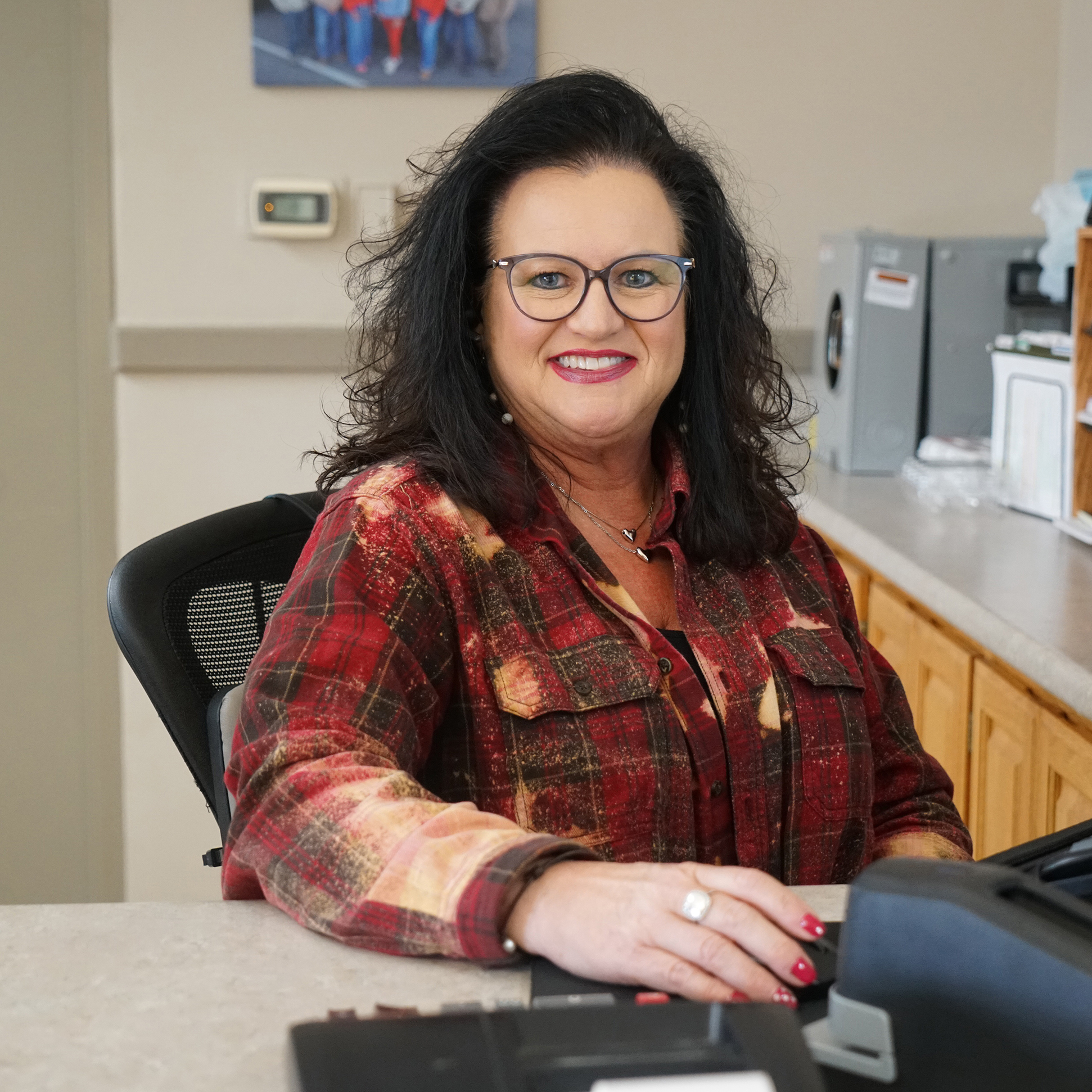 Smiling woman seated at a customer service desk in an office, wearing glasses and a red plaid shirt