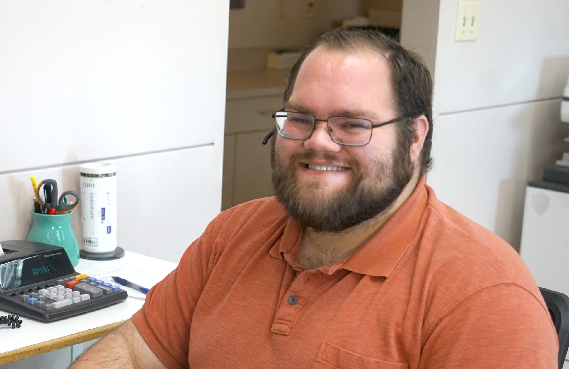 Smiling man seated at an office desk, wearing an orange short-sleeve polo shirt and glasses, looking toward the camera
