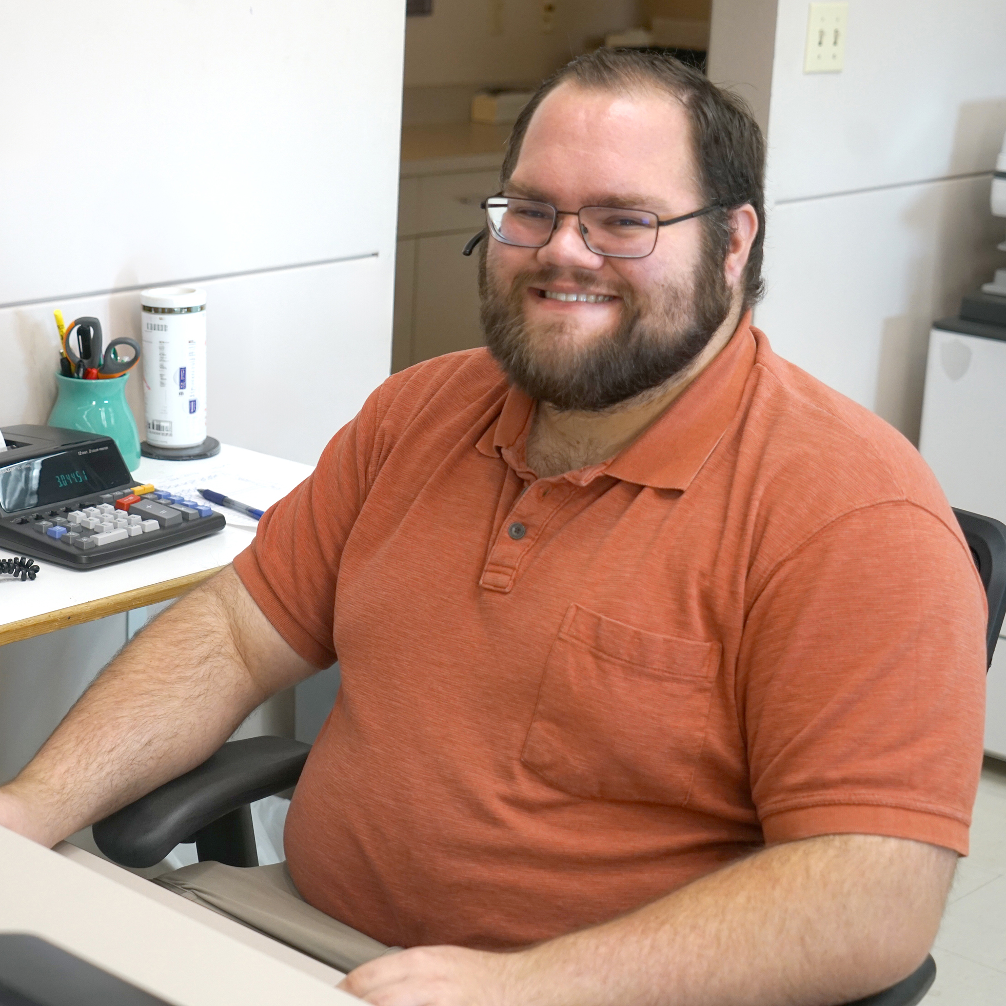 Smiling man seated at an office desk, wearing an orange short-sleeve polo shirt and glasses, looking toward the camera