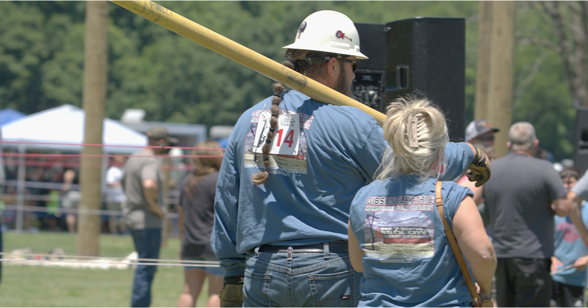 An electric lineworker and his wife stand side by side at a lineman rodeo, seen from behind as he carries a long insulated tool over his shoulder and she gestures toward the competition area.