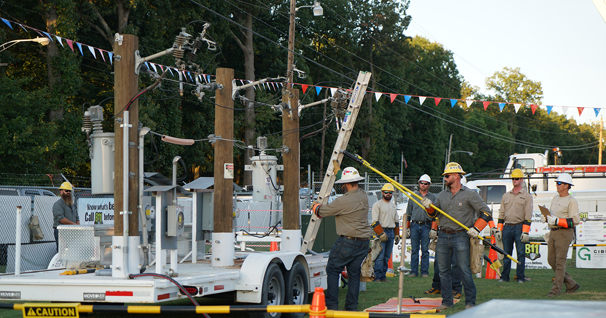 Lineworkers participate in a safety demonstration showing proper ladder placement and distance from energized overhead power lines.