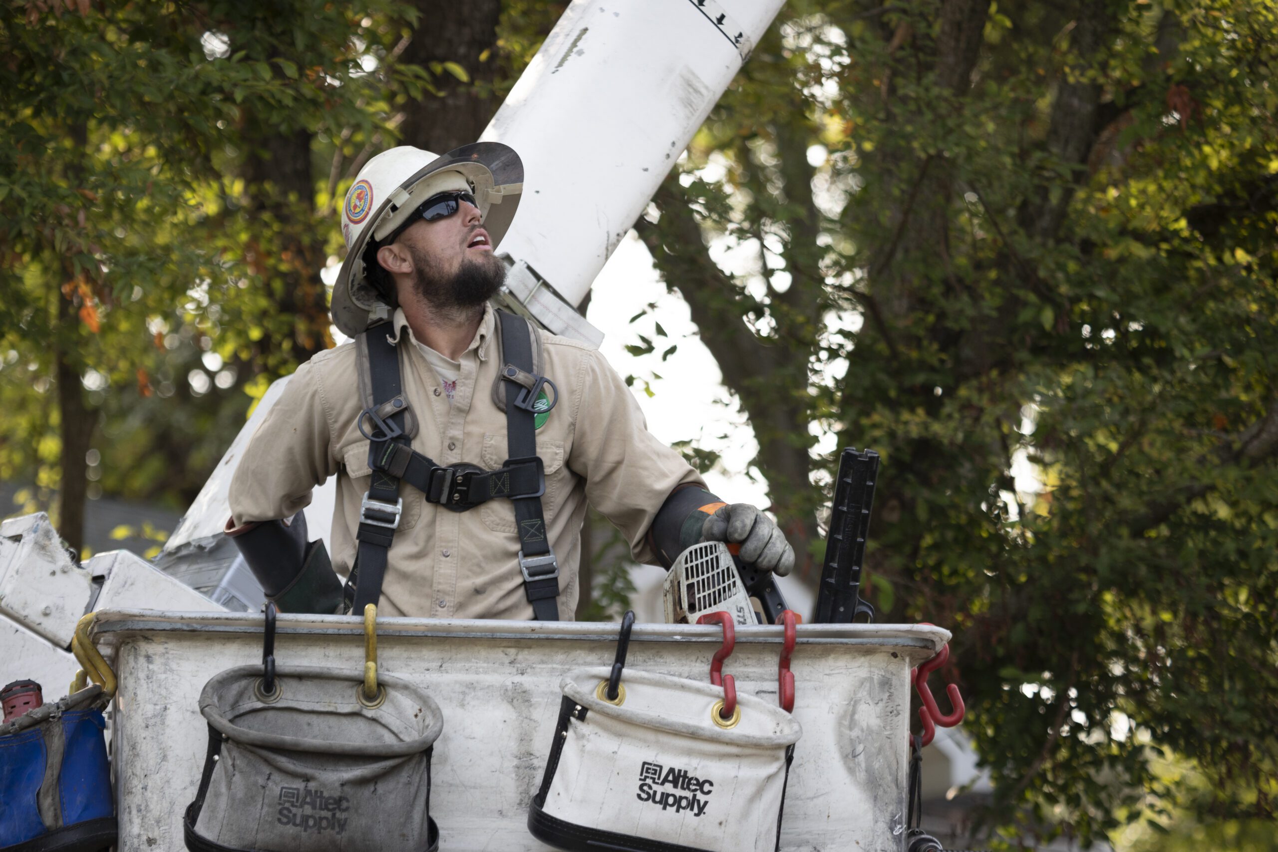 Utility worker wearing a hard hat and safety harness stands in a bucket lift, looking up while working near power lines, with tool bags hanging from the bucket and trees in the background.