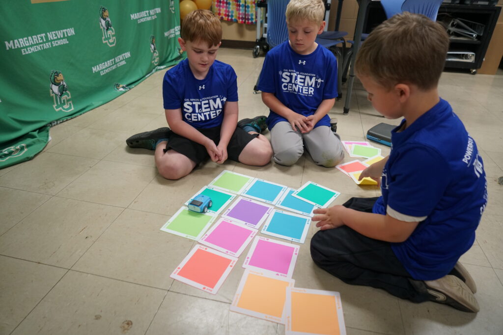 Three students wearing STEM Center shirts sit on the floor and use color-coded cards to program a screen-free coding robot in the Ripken Foundation STEM Center.