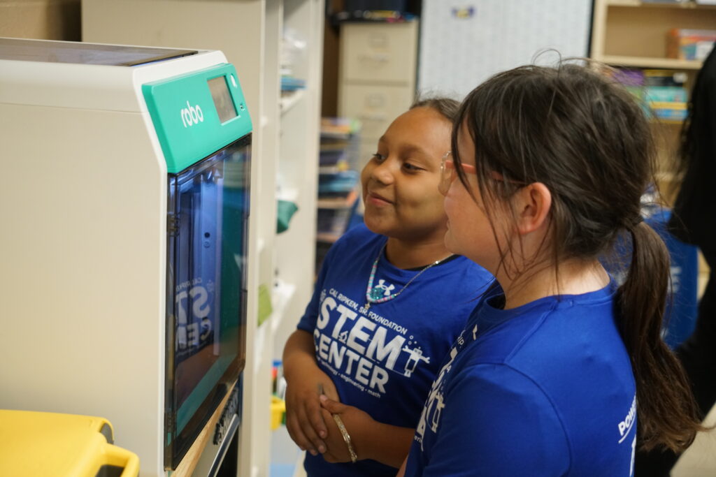 Two students wearing STEM Center shirts watch a 3D printer as it creates a keychain in the Ripken Foundation STEM Center.