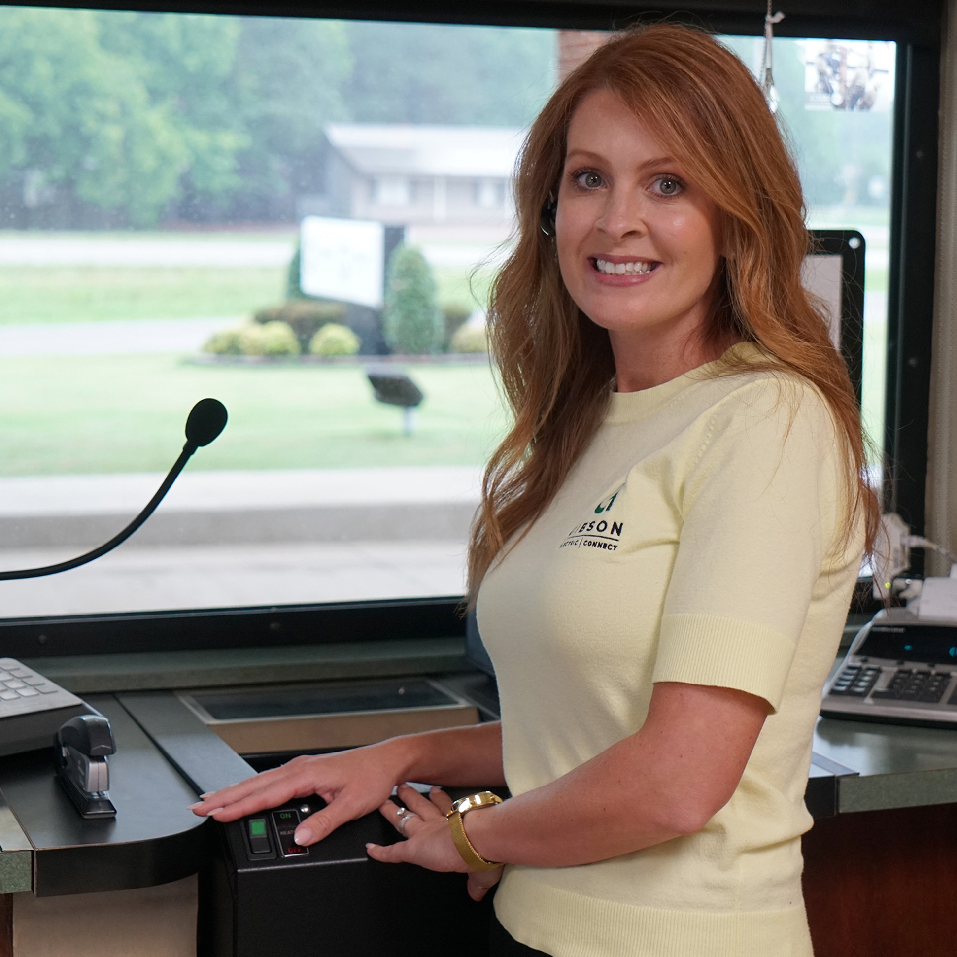 Employee at a service counter wearing a headset, operating a control panel and microphone, with a window and landscaped outdoor area in the background.