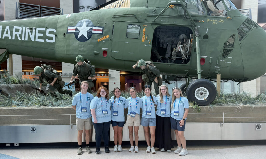 Seven Washington Youth Tour participants wearing light blue event T-shirts stand in front of a Vietnam-era Marine Corps helicopter exhibit featuring life-size military figures at the National Museum of the Marine Corps.
