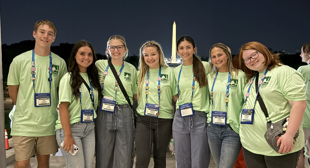 Seven Washington Youth Tour participants wearing matching light green T-shirts and conference badges pose for a group photo at night, with the illuminated Washington Monument visible in the background.