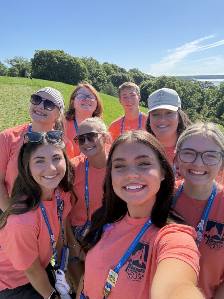  Seven Washington Youth Tour participants and their chaperone wearing matching coral event T-shirts gather for a selfie outdoors on a sunny day, with rolling green hills, trees and a distant water view in the background.