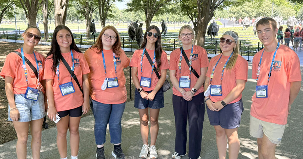 Seven young people wearing matching coral-colored event T-shirts and conference badges stand together on a shaded walkway at a memorial site. Rows of trees, visitors, and dark statues are visible in the background.