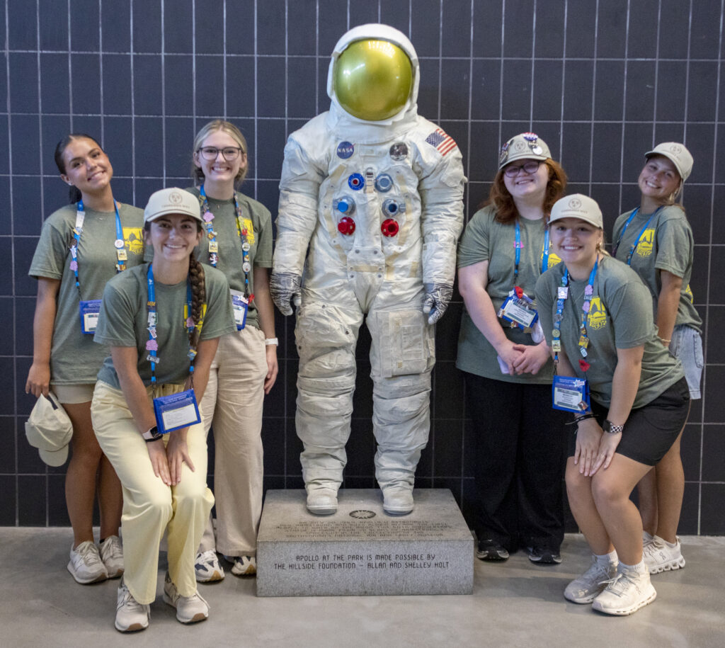 Six Washington Youth Tour participants wearing green event T-shirts, conference badges and Tennessee Youth Tour caps pose around a NASA astronaut suit exhibit displayed against a dark tiled wall.