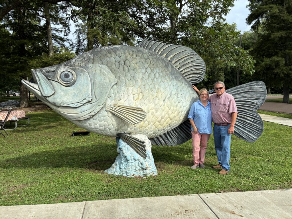 Two people stand beside a giant crappie statue in a park-like setting with large trees and green grass in the background. The oversized fish sculpture towers over the visitors and is mounted on a pedestal designed to resemble water.